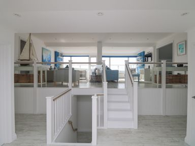 A split-level room with a staircase leading to an upper living area with sofas tables shelves and large windows at St Ives View in Porthtowan