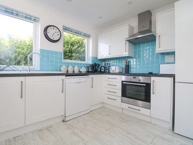 A kitchen with white cabinets blue tiled backsplash dishwasher oven and windows with garden views at St Ives View in Porthtowan