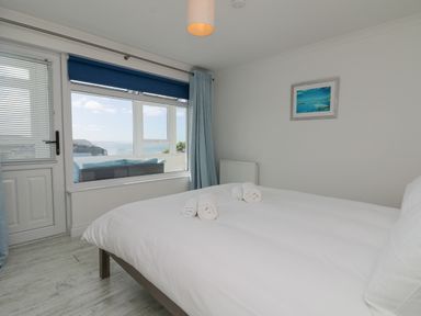 A bedroom with a bed and rolled towels near a window showing an outdoor seating area and sea view at St Ives View in Porthtowan