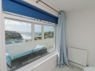 A room with a large window showing a view of the ocean and cliffs with a wicker sofa outside and blue curtains at St Ives View in Porthtowan
