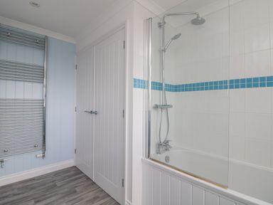 A bathroom with a bathtub with shower and blue tiled border near a white double door and chrome towel radiator at St Ives View in Porthtowan