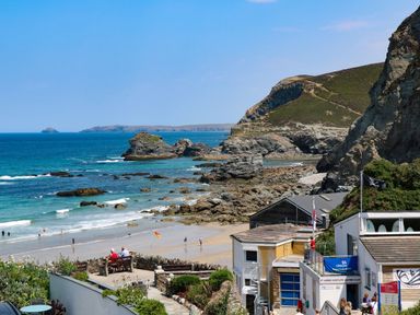 A rocky beach with people walking near the shore and buildings with signs by the coast at St Ives View in Porthtowan