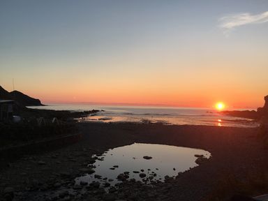 A rocky beach with a small pool of water at sunset with the sun near the horizon at Oaklands Near Boscastle