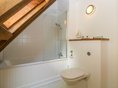 A bathroom with a bathtub shower area under a sloped wooden ceiling and a toilet next to a shelf with decorative items at Oaklands Near Boscastle