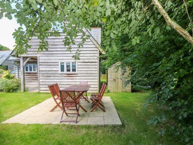 An outdoor patio with a wooden table and six chairs next to a wooden shed and house at Oaklands Near Boscastle
