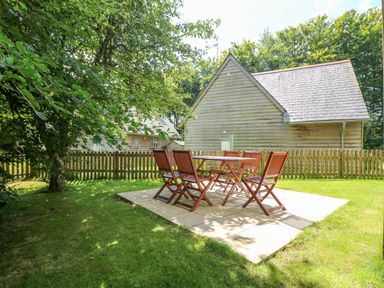 A wooden outdoor table with six chairs on a patio in a backyard with a wooden fence and houses near trees at Oaklands Near Boscastle