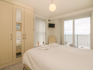 A bedroom with a bed towels wardrobe chair and a TV next to a balcony with sea view at Seasiders in Ilfracombe