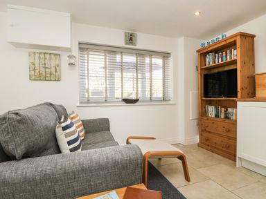 A sitting area with a gray sofa and striped cushions next to a window with blinds and a wooden media cabinet at Seascape in Porthcurno