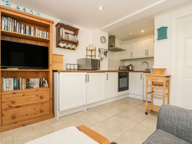 A kitchen area with white cabinets and wooden countertops next to a wooden shelving unit with a television and books at Seascape in Porthcurno