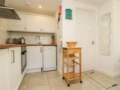 A kitchen with white cabinets a dishwasher oven kettle and wooden trolley with bowl at Seascape in Porthcurno