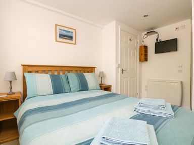 A bedroom with a double bed striped bedding and folded towels two bedside tables with lamps a wall-mounted TV and a door at Seascape in Porthcurno