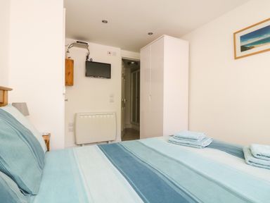 A bedroom with a striped blue bedspread a white wardrobe a wall-mounted TV and a framed picture at Seascape in Porthcurno