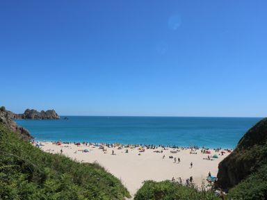 A sandy beach with people sunbathing and swimming by the sea with rocky cliffs on the sides at Seascape in Porthcurno