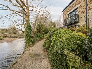 A gravel pathway along a river with bushes on one side and a stone building with balconies on the other at Riverview in Lostwithiel