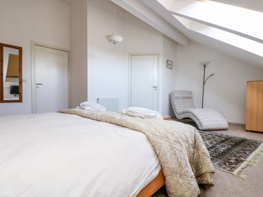 A bedroom with a bed, a patterned chaise lounge, a floor lamp, a wooden dresser, a mirror, and two doors at Riverview in Lostwithiel