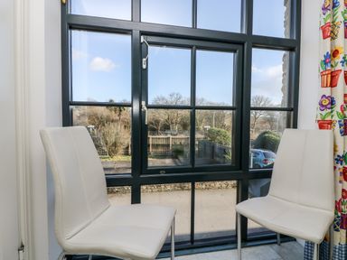 Two white chairs in front of a large window with a black frame overlooking a road and trees at Riverview in Lostwithiel