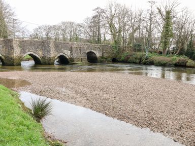 A stone bridge over a river with a pebbled riverbank and trees in the background at Riverview in Lostwithiel