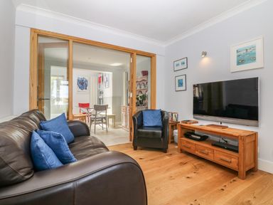 A living room with a brown leather sofa and armchair, blue cushions, wooden TV stand with a flat screen TV and glass sliding doors leading to a dining area at Tides in Padstow