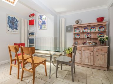 A dining area with a glass table wooden and gray chairs a wooden cabinet with colorful plates and flowers at Tides Padstow