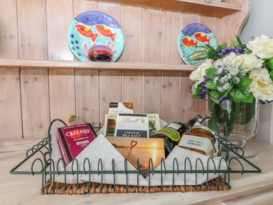 A wire basket with coffee pods chocolates marmalade and a small bottle next to a glass vase with flowers on a wooden surface at Tides in Padstow