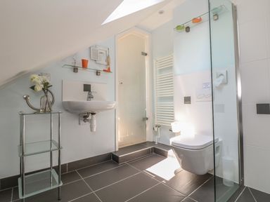 A bathroom with a wall-mounted sink and toilet a glass shelf with a cup and decoration a small glass table with flowers and a radiator at Tides in Padstow