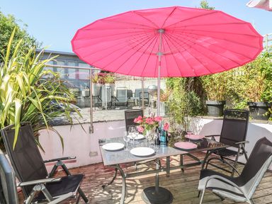 An outdoor dining area with a glass table and black chairs under a pink umbrella at Tides in Padstow