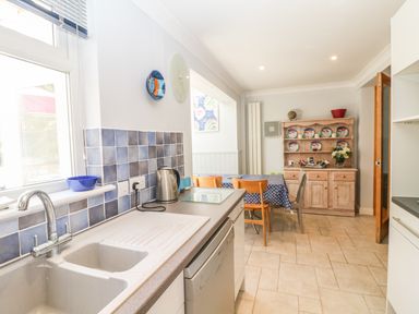 A kitchen with a sink and countertop next to a dining area with a table and chairs and a wooden cabinet displaying plates at Tides in Padstow