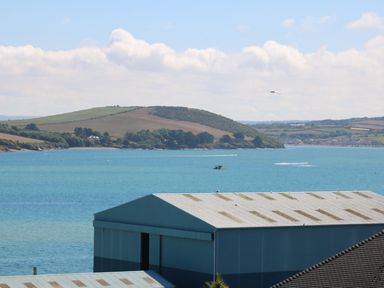 A body of water with boats and a large blue industrial building in the foreground and a green hill in the distance at Tides in Padstow