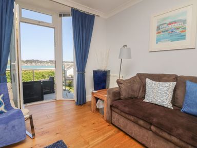 A living room with a brown sofa blue curtains and a door opening to a balcony overlooking water at Tides in Padstow