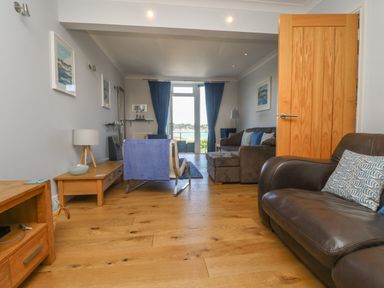 A living room with wooden flooring sofas a chair tables blue curtains and a door with a waterfront view at Tides in Padstow