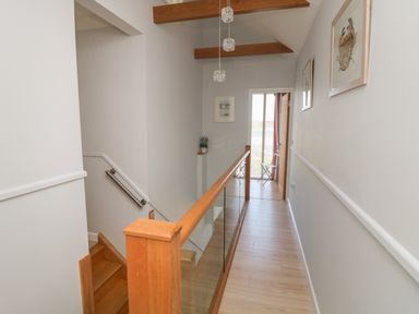 A hallway with wooden stairs and railing with glass panels leading to a door with a view outside at Tides in Padstow