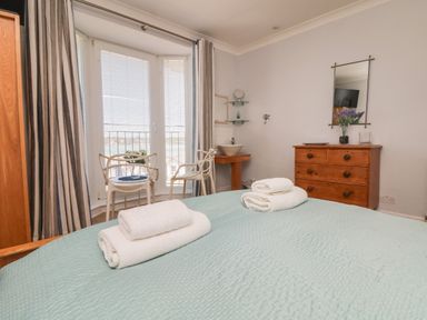 A bedroom with a bed, two rolled towels, two chairs by windows with blinds, a wooden dresser with a mirror and flowers at Tides in Padstow