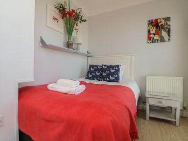 A bedroom with a bed covered by a red blanket and two folded white towels on top a white bedside table with a drawer a shelf with a vase of flowers and a lamp and framed pictures on the wall at Tides in Padstow