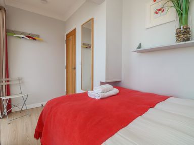 A bedroom with a bed covered by a red blanket and folded towels on top a wooden door a full-length mirror and a chair at Tides in Padstow