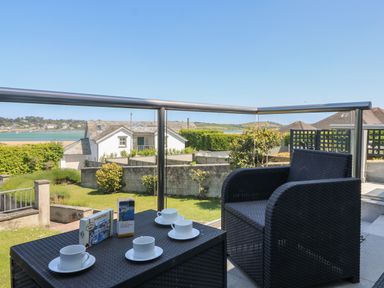 A balcony with a black woven table and chair and four white cups with saucers overlooking a garden and houses at Tides Padstow