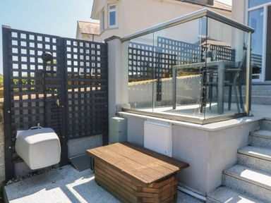 An outdoor area with a tiled shower head, wooden storage bench, stairs, and a glass railing with a table and chairs behind it at Tides in Padstow