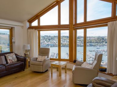 A sitting area with two armchairs a sofa and a wooden table in front of large windows overlooking a marina at Totherside in Kingswear