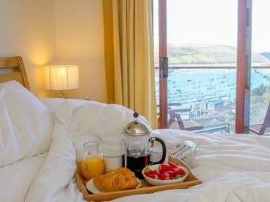 A bedroom with a wooden bed and white bedding with breakfast tray including croissants strawberries orange juice and coffee near a window with a view of a harbor at Totherside in Kingswear