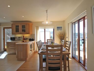 A dining area with a wooden table set for six next to large sliding glass doors and a kitchen with wooden cabinets at Totherside in Kingswear