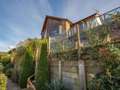 A stone house with large windows and glass balcony above a wooden retaining wall with plants and trees at Totherside in Kingswear