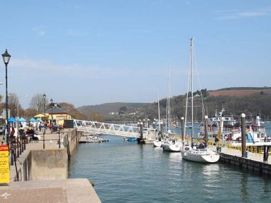 A harbor with boats docked along a pier and people walking on a waterfront promenade at Totherside in Kingswear