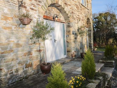 Stone building entrance with potted plants hanging baskets and yellow flowers at Lost Barn in Watergate Bay