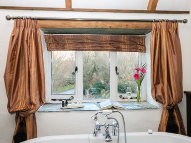 A bathroom window with brown curtains a book a glass of champagne and flowers at Lost Barn in Watergate Bay