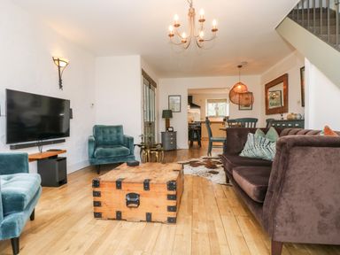 A living room with two blue armchairs a brown sofa a wooden trunk coffee table a wall-mounted television and a dining area at Lost Barn in Watergate Bay