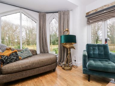 A living room with a curved brown sofa and a green armchair near windows with gray curtains and a flamingo lamp at Lost Barn in Watergate Bay