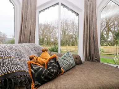 A brown sofa with patterned pillows and a blanket near large windows with curtains showing a fenced garden outside at Lost Barn in Watergate Bay