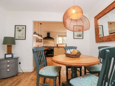 A dining area with wooden table and four upholstered chairs next to a gray side table with a lamp and a kitchen in the background at Lost Barn in Watergate Bay