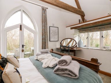 A bedroom with a wooden bed, folded towels, bathrobes, a dressing table with a mirror and chair, an arched window, and a smaller window with a vase at Lost Barn in Watergate Bay