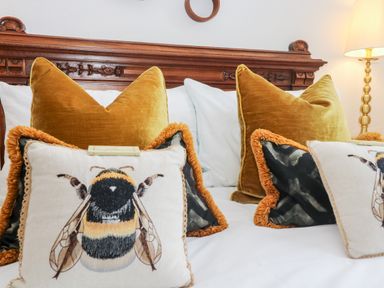 A bed with decorative pillows including bee print pillows in front of large mustard cushions and a wooden headboard with a lamp nearby at Lost Barn in Watergate Bay