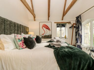 A bedroom with a bed featuring floral and dark pillows a green blanket folded towels a flamingo painting exposed wooden beams and windows with dark curtains at Lost Barn Watergate Bay